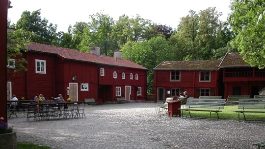 Röda gamla trähus vid ett torg av kullersten. På torget ses även människor och några bänkar.