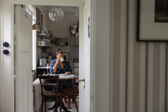 En kvinna i blå-rutig klänning sitter vid ett runt bord i ett mysigt kök och dricker ur en kopp. Bordet är omgivet av mörka trästolar, och köket är inrett med rustika detaljer.